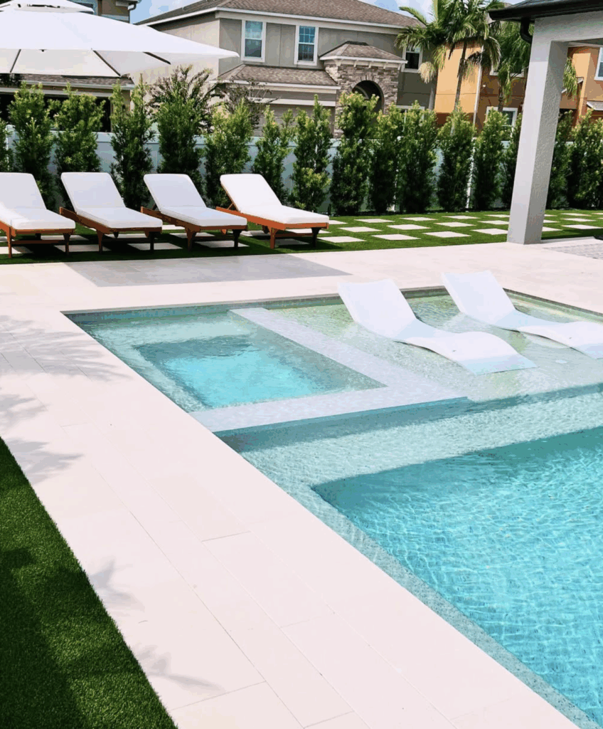 A modern backyard with a pool and built-in spa, three white lounge chairs in the water, and four poolside loungers on a grass lawn.